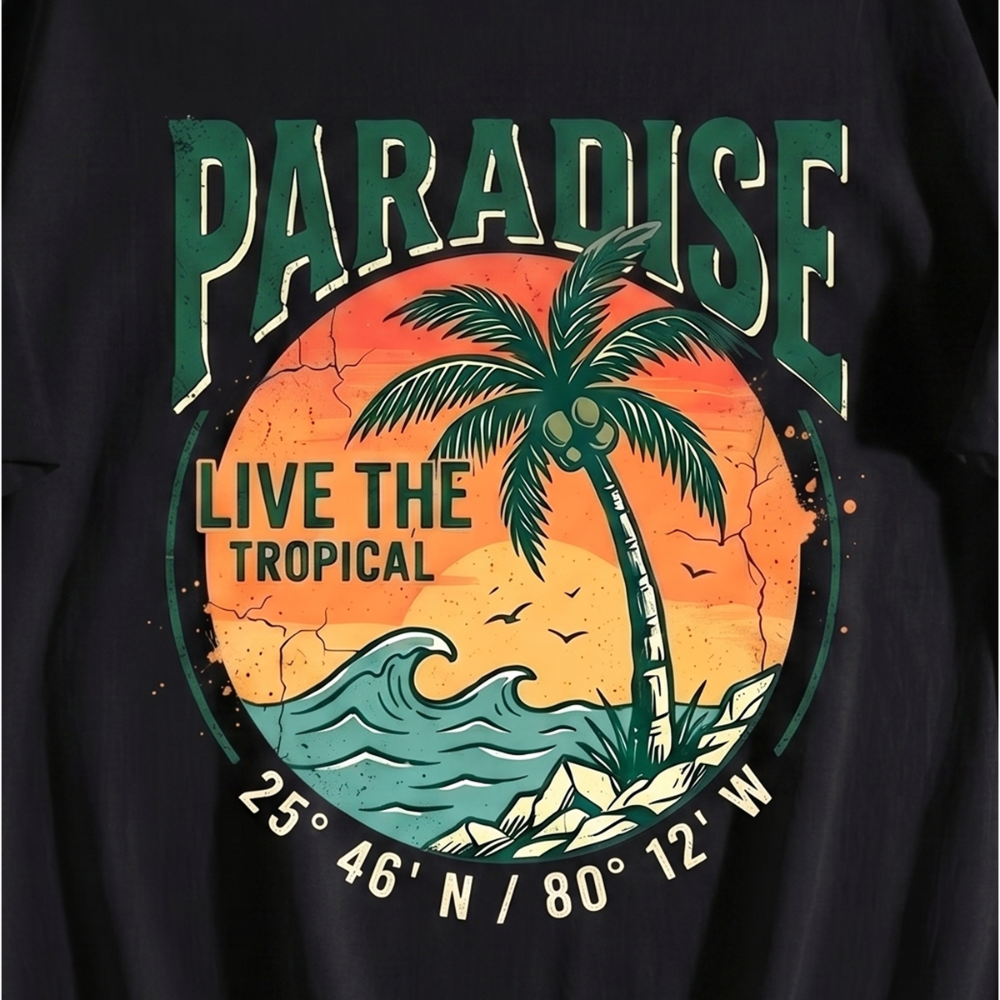 A close-up of a black graphic t-shirt featuring a retro tropical design with a palm tree, ocean wave, and the text 'PARADISE LIVE THE TROPICAL' along with coordinates.