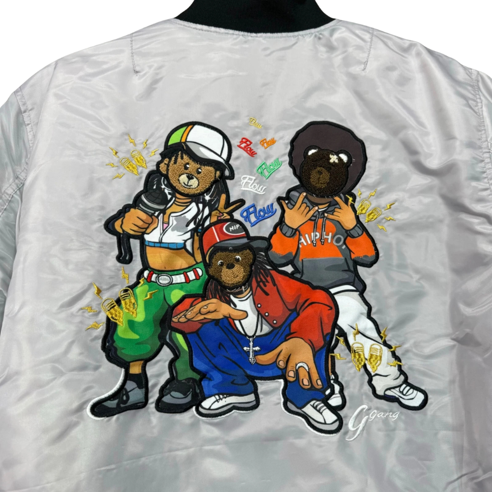 Close-up of the back of a shiny light gray bomber jacket with a large colorful cartoon graphic of three teddy bears in streetwear, including one wearing a green bag and the other in a red jacket. The collar is black.