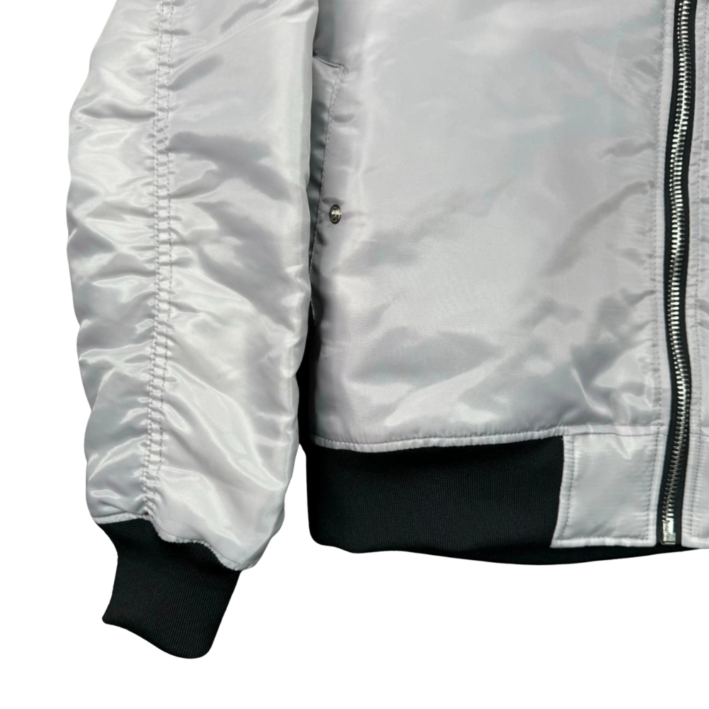 Close-up of a shiny light gray bomber jacket with black ribbed cuffs, hem, and a black zipper, shown on a white background.