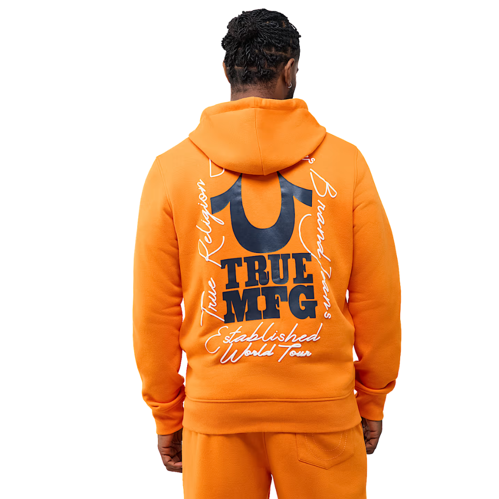 A man viewed from behind wearing a bright orange hoodie and matching sweatpants, featuring a large navy and white graphic print on the back with the text 'TRUE MFG' and 'True Religion Established World Tour'.