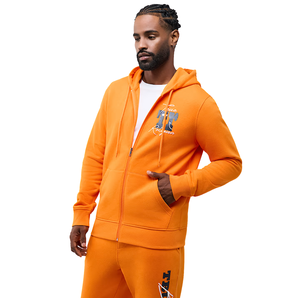 A man modeling a bright orange zip-up hoodie and matching sweatpants set against a plain white background. The hoodie features a graphic print on the left chest and a full front zipper.